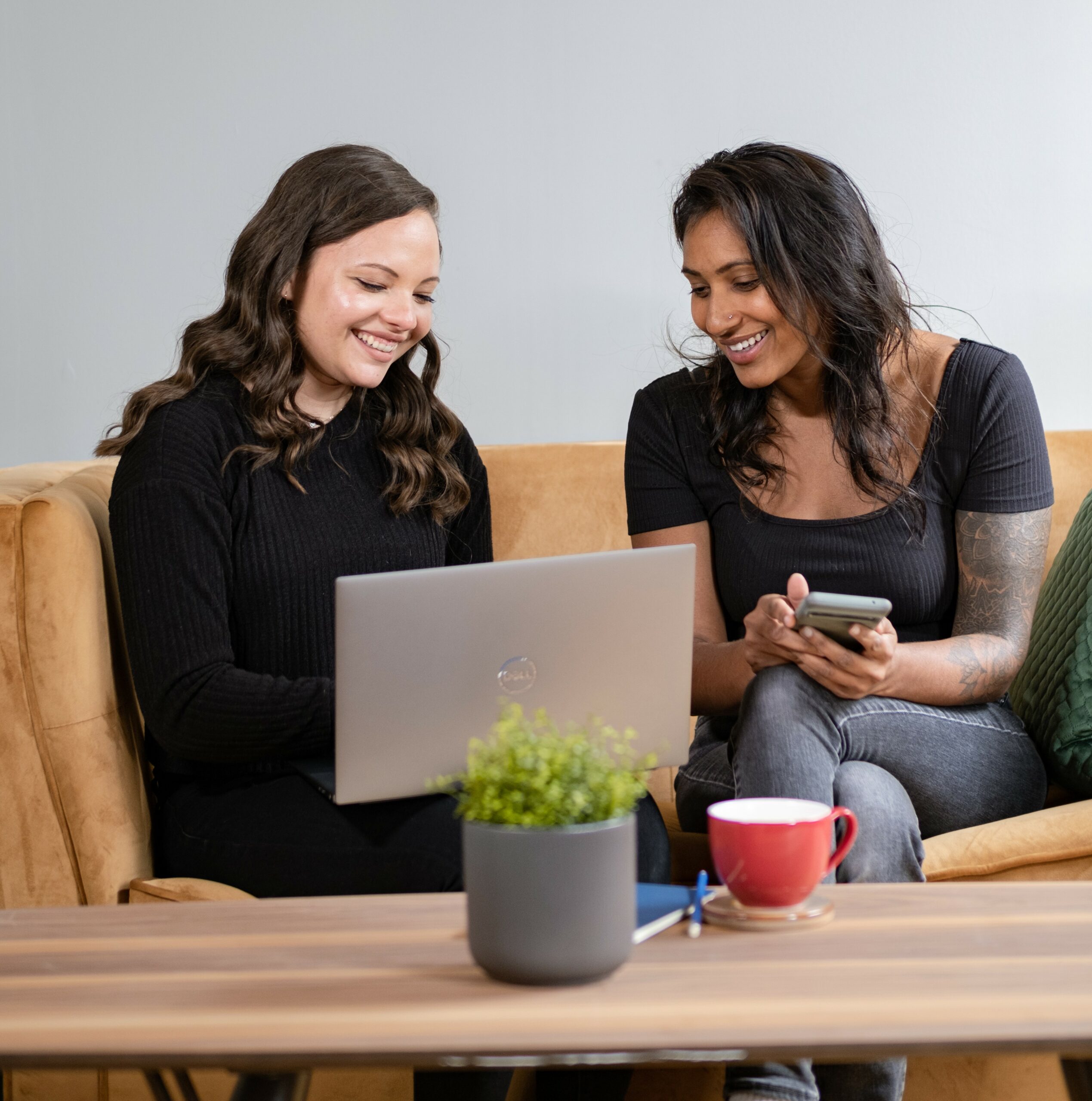 Two women sitting on a beige couch smiling at a laptop